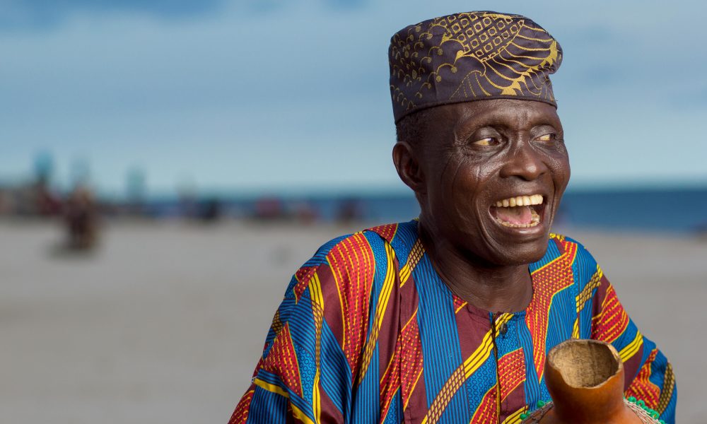 Elderly man joyfully smiling wearing Yoruba attire and cap on Lagos beach.