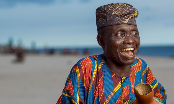 Elderly man joyfully smiling wearing Yoruba attire and cap on Lagos beach.